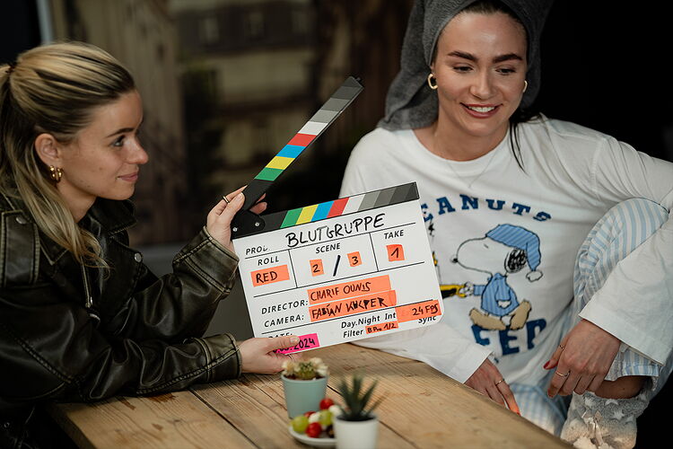 Zwei Darstellerinnen an einem Tisch, davor die Filmklappe zur Produktion Blutgruppe